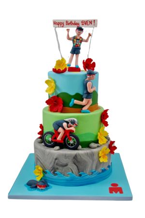 Un grand gâteau d'anniversaire à trois étages sur le thème du triathlon Ironman, représentant un nageur dans l'eau, un cycliste sur un chemin rocheux, un coureur et un athlète passant la ligne d'arrivée.