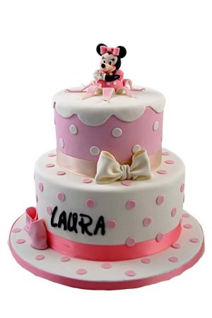 Une pièce montée d'anniversaire à deux étages en pâte à sucre blanche à pois roses et rose à pois blancs, ornée de nœuds en sucre et surmontée d'une figurine 3D de Minnie Mouse assise au sommet.