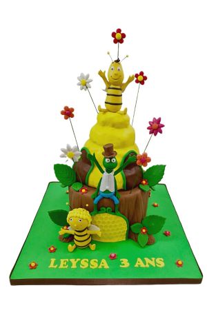 Pièce montée d'anniversaire sculptée Maya l'Abeille en forme de souche d'arbre et ruche jaune avec figurines de Flip la sauterelle, Willy et Maya entourés de fleurs sur tiges réalisée par The French Cake Company